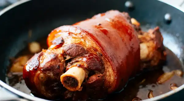Un magnifique jarret de porc au four, la peau dorée et brillante, mijote dans sa sauce savoureuse avec des gousses d'ail.