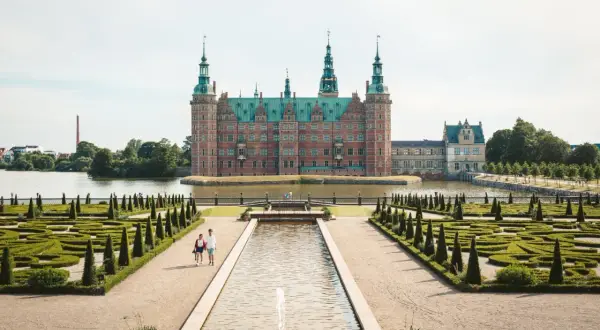 Ett imponerande slott i Danmark, omgivet av vatten och med en vacker barockträdgård. Ett par promenerar längs en damm framför slottet.