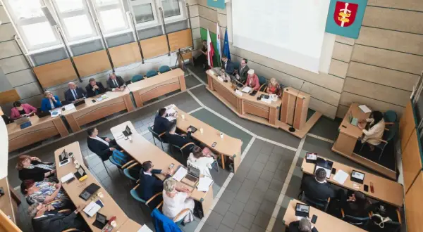 Obrady rady miasta, podczas których odbywa się udzielenie absolutorium. Ludzie siedzą przy stołach, niektórzy pracują na laptopach.