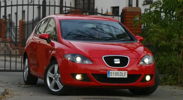 Czerwony Seat Leon 2 z włączonymi światłami, zaparkowany przed bramą.