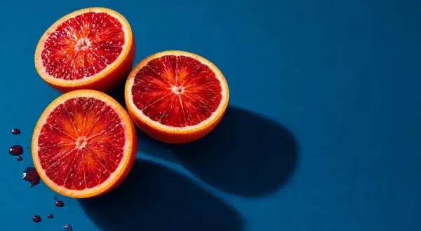 Vibrant fruit photography: three blood oranges halved, revealing their deep red flesh, with a few juice droplets on a deep blue background.