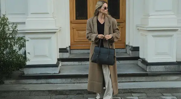 En kvinna med solglasögon bär en elegant brun kappa och håller en stilren vero moda väska.