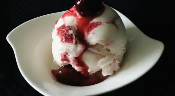 Ein Löffel cremiges Sauereis mit Kirschsauce und einer ganzen Kirsche obendrauf, serviert in einer weißen Schale.