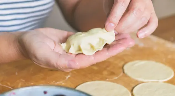 Dłonie formują pieroga z ciasta na pierogi bez jajka. Na blacie leżą okrągłe placki ciasta.