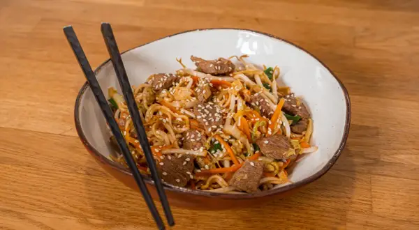 Makaron Chow Mein z wołowiną, warzywami i sezamem w misce z pałeczkami.