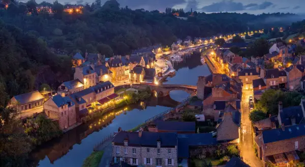 Widok na urokliwe francuskie miasteczko nad rzeką o zmierzchu, z oświetlonymi budynkami i mostem.