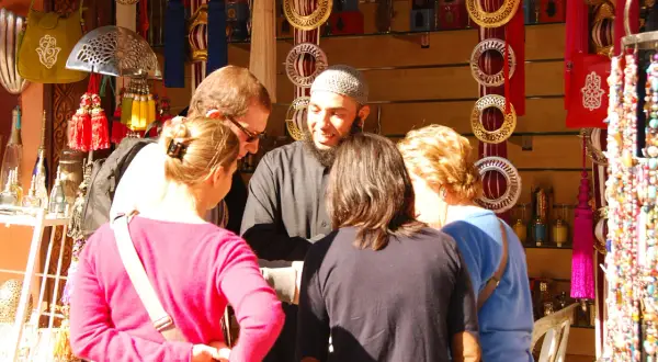 Un groupe de touristes discute avec un vendeur dans un souk de Marrakech, entouré de lanternes, de bijoux et de textiles colorés.