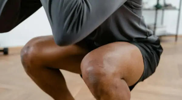 Ein Mann macht Kniebeugen, seine Knie sind gebeugt. Er hat leichte Narben auf dem rechten Knie, die auf frühere Schmerzen in der Kniekehle hindeuten könnten.
