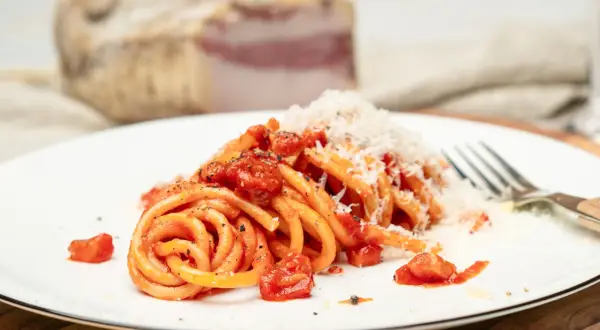 Ein Teller Pasta all'amatriciana, mit Tomaten und geriebenem Käse bestreut. Im Hintergrund ein Stück Guanciale, die Zutat für das authentische amatriciana rezept.