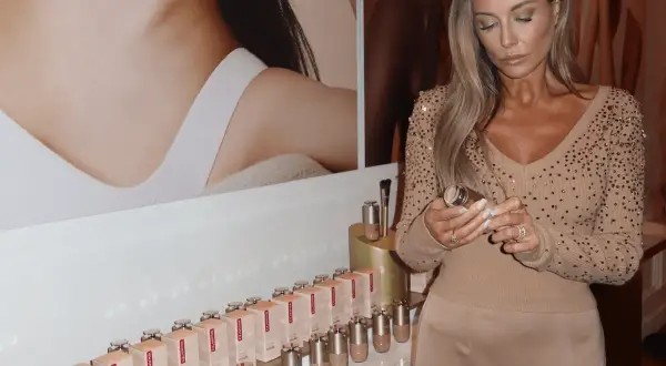 Kobieta z długimi blond włosami trzyma buteleczkę Clarins Double Serum Foundation. Na półce stoją dziesiątki opakowań tego podkładu.