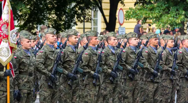 Żołnierze w mundurach defilują z karabinami. W tle sztandar i drzewa. Kategoria e wojsko.