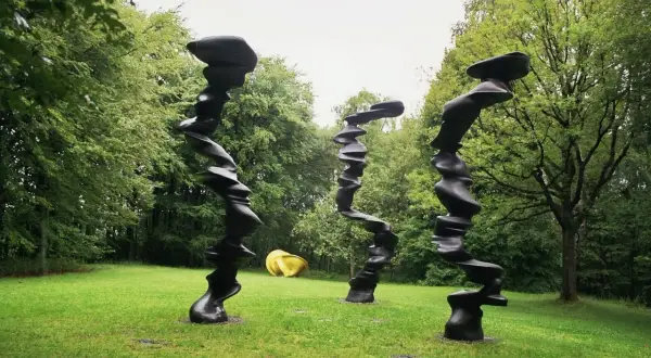 Drei schwarze, verdrehte Skulpturen stehen im **Skulpturenpark Wuppertal** auf einer grünen Wiese vor Bäumen. Eine gelbe Skulptur liegt im Hintergrund.