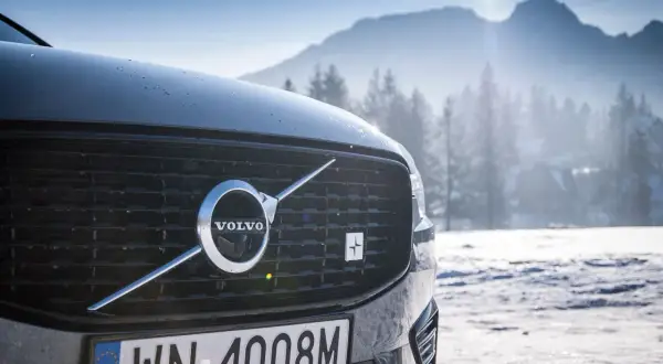 Szary Volvo XC60 Polestar z emblematem Polestar na grillu, zaparkowany na tle ośnieżonych gór i lasu.