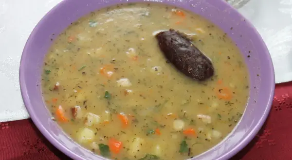 Gęsta zupa fasolowa z puszki, z kawałkami warzyw i ciemną kiełbaską, podana w fioletowej misce.
