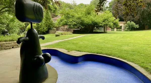 Eine abstrakte schwarze Skulptur steht neben einem blauen Mosaikbecken in einem **Skulpturenpark**. Im Hintergrund ein gepflegter Rasen und Bäume.