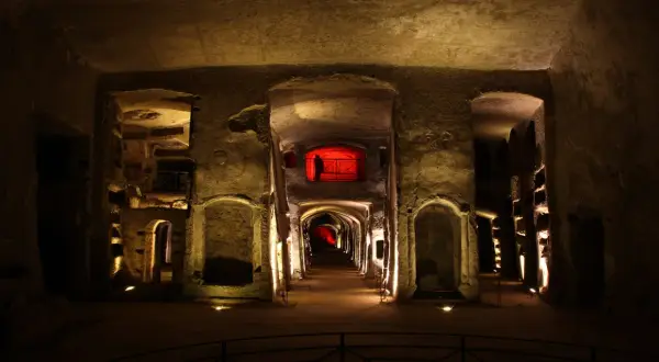 Corridoio sotterraneo illuminato, con nicchie scavate nella roccia e un'apertura rossa in lontananza, evocando l'atmosfera della tomba di San Gennaro.