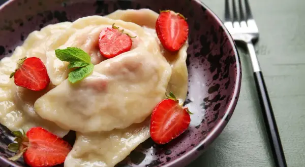 Pyszne pierogi z truskawkami, udekorowane świeżymi owocami i miętą, czekają w misce na widelcu.