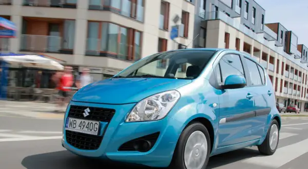 Błękitny Suzuki Splash jedzie ulicą miasta. W tle nowoczesne budynki i ludzie.
