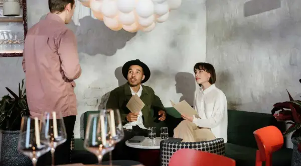Kelner w różowej koszuli rozmawia z parą przy stoliku. Zastanawiają się, ile zarabia kelner, przeglądając menu.