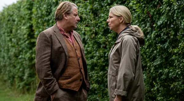 Två personer står och pratar intensivt framför en grön häck. Mannen bär kavaj och väst, kvinnan en jacka. En scen från en dansk kriminalserie på TV4.