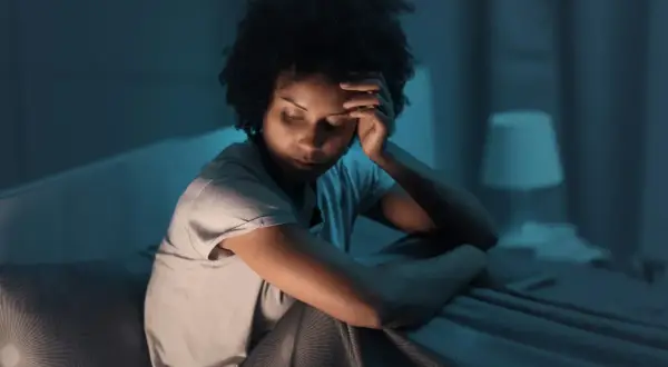 A woman sits in bed, head in hand, eyes closed, appearing to be experiencing fragmented sleep causes.