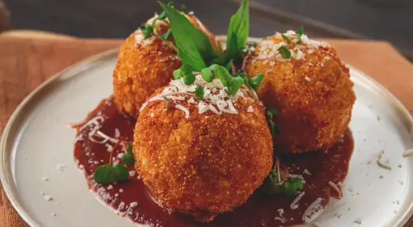 Trois arancini, spécialité italienne, dorés et croustillants, servis sur une sauce rouge avec des herbes fraîches et du parmesan râpé.