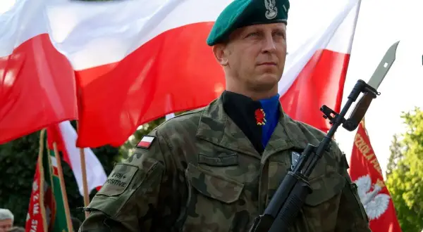 Dumnie stoi dowódca w zielonym beretcie, z karabinem i polską flagą w tle.