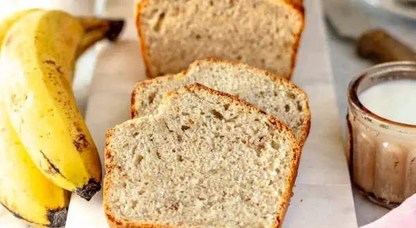 Fetta di banana bread senza uova, soffice e dorata, accanto a banane mature e un bicchiere di latte.