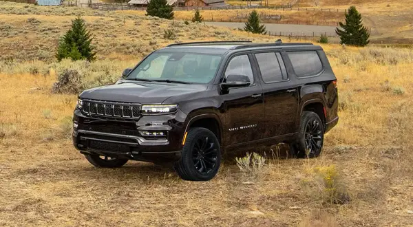 Ciemny Jeep Grand Wagoneer na tle wiejskiego krajobrazu z budynkami i drzewami.