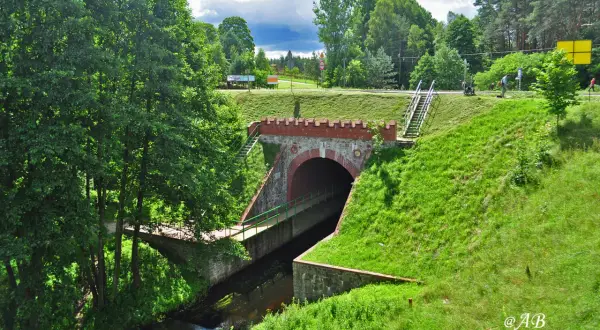 Kamienny tunel z ceglanym łukiem, przez który przepływa strumień. Nad nim zielone, trawiaste zbocza i drzewa. W oddali widać drogę i ludzi. To miejsce przypomina stare akwedukty.