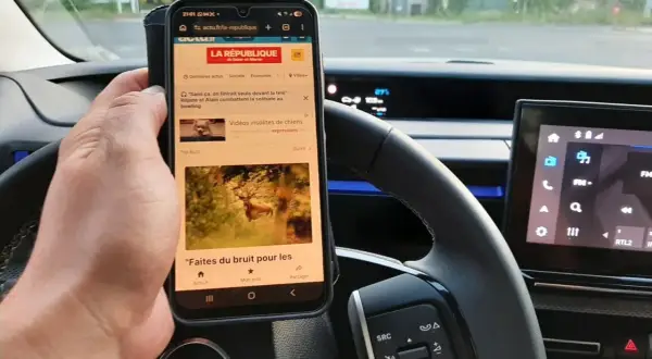 Main tenant un téléphone au volant, risque d'amende. L'écran affiche des articles d'actualité, un danger pour la conduite.