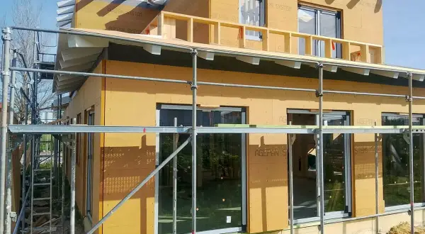 Casa en construcción con paredes exteriores prefabricadas de madera, andamios y cielo azul.