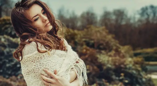 Una giovane donna con capelli castani, avvolta in una sciarpa di pizzo, sembra pensare a se stessa in un parco autunnale.