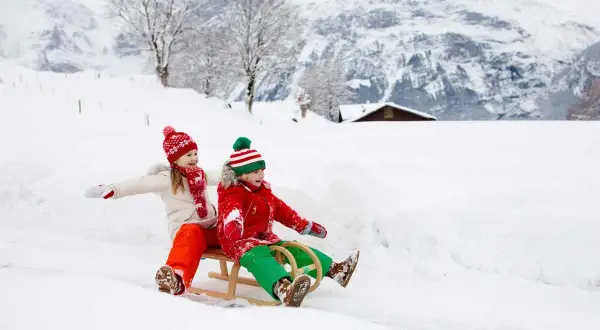Zwei Kinder rodeln fröhlich durch den Schnee. Ein idyllisches Bild vom Winter in der Schweiz mit verschneiten Bergen und einem kleinen Haus im Hintergrund.