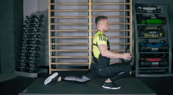 Mężczyzna wykonuje ćwiczenia mobilizacyjne z kettlebell, pogłębiając wykrok. W tle stojaki z hantlami i fitbagami.