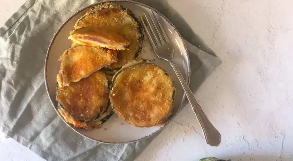 Cordon bleu di melanzane impanati e fritti, ripieni di formaggio filante e prosciutto, serviti su un piatto con una forchetta.
