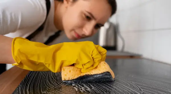 Kobieta w żółtej rękawiczce czyści płytę indukcyjną gąbką z pianą. Dowiedz się, czym czyścić płytę indukcyjną, by lśniła czystością.