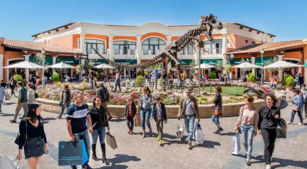 Un T-Rex scheletrico domina il centro del campo rom Castel Romano, circondato da negozi e gente che fa shopping.