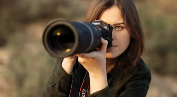 Kobieta z aparatem Canon EOS R7 uczy się, jak robić zdjęcia. Skupiona na kadrze, z obiektywem skierowanym w stronę widza.