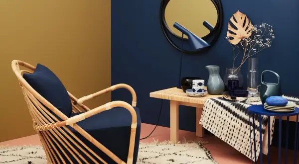 Ein Rattansessel mit dunkelblauem Kissen steht auf einem flauschigen Teppich. Daneben ein Tisch mit Deko. Welche Farbe passt zu Gold? Dunkelblau und Senfgelb harmonieren perfekt.