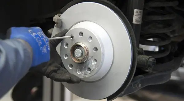 Mechanik w niebieskiej rękawicy dokręca śrubę podczas wymiany tarcz hamulcowych. Widać nowy, lśniący dysk hamulcowy i sprężynę amortyzatora.