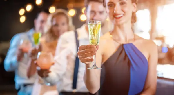Uśmiechnięta kobieta trzyma kieliszek z drinkiem z prosecco i wódką, w tle bawią się inni goście.