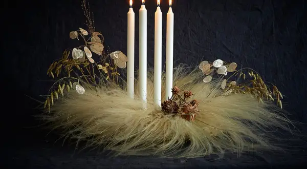 Ausgefallener Adventskranz Trend: Vier weiße Kerzen auf einem Bett aus Federn und Gräsern, geschmückt mit Lunaria und Zapfen.