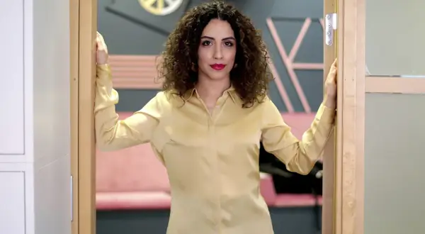 Helya Riazat, med lockigt mörkt hår och rött läppstift, står i en dörröppning i en guldig blus.