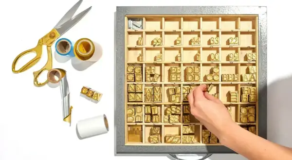 A hand selects brass letter stamps from a tray, ready to create a monogram. Gold scissors and foil rolls are nearby.