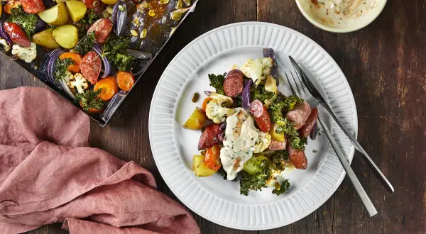 En tallrik med ugnsrostade grönsaker och korv, ett perfekt potatis recept. Bredvid syns en ugnsplåt med samma läckra innehåll.