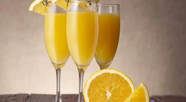 Tre glas med en gyllene drink med cava, garnerade med apelsinskivor och rosmarin. En apelsin och lavendel på träbordet.