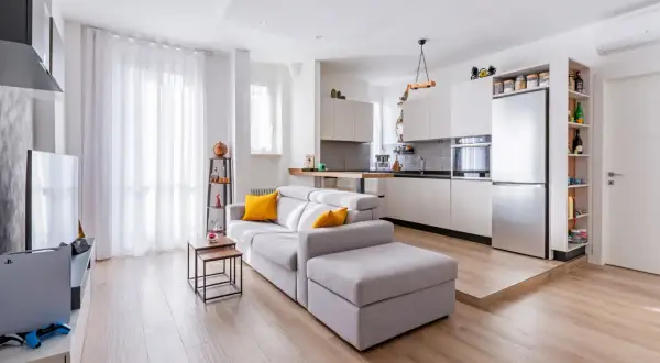 Un ampio open space moderno con divano grigio, cucina a vista e pavimento in legno chiaro.