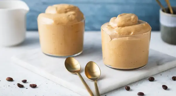 Due bicchieri di cremosa schiuma di caffè, perfetta per la tua ricetta crema di caffè senza panna. Due cucchiaini dorati e chicchi di caffè completano la scena.
