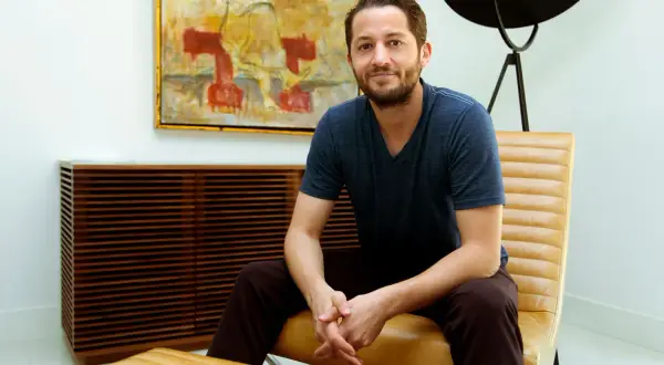 Ian Kirkpatrick, a man with a beard and a blue t-shirt, sits in a tan leather chair in a modern room with abstract art.